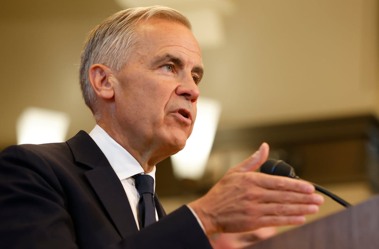 Man with white and grey hair wears a black suit with a white shirt and black tie while speaking into a microphone at a podium