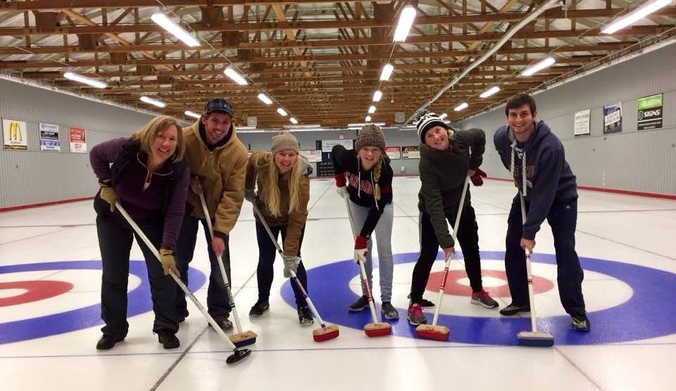 default-80 Two men and four women with sweeping brooms stand on the painted circles of a curling rink.