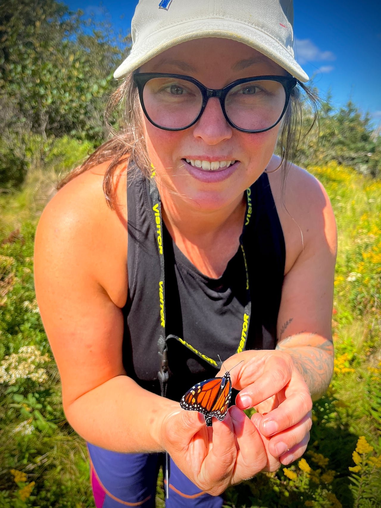 default-90 A woman in glasses and a white cap holds a monarch butterfly with a small tag on its thorax in a field of yellow wildflowers.